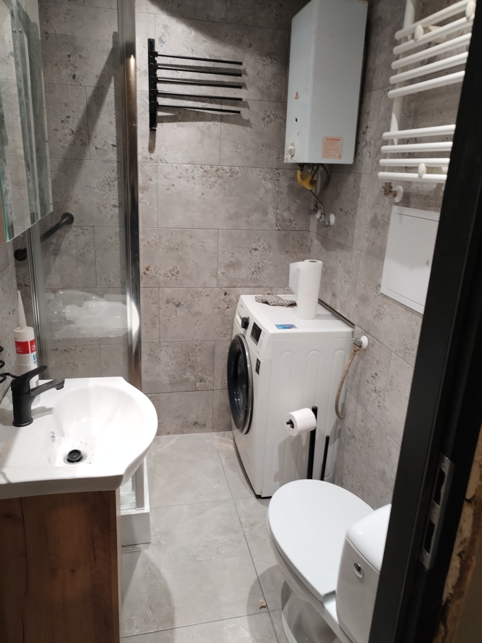 Wyremontowana łazienka w szarościach z pralką, umywalką z szafką z drewnopodobnym wykończeniem, grzejnikiem drabinkowym i bojlerem.