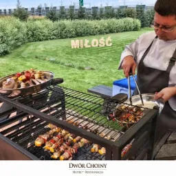 Kucharz w fartuchu przekłada szaszłyki z grilla do metalowej miski; w tle trawnik z napisem 'MIŁOŚĆ' z drewnianych liter.
