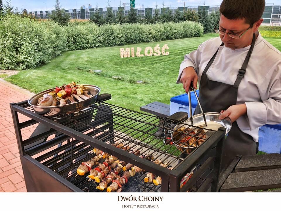 Kucharz w fartuchu przekłada szaszłyki z grilla do metalowej miski; w tle trawnik z napisem 'MIŁOŚĆ' z drewnianych liter.