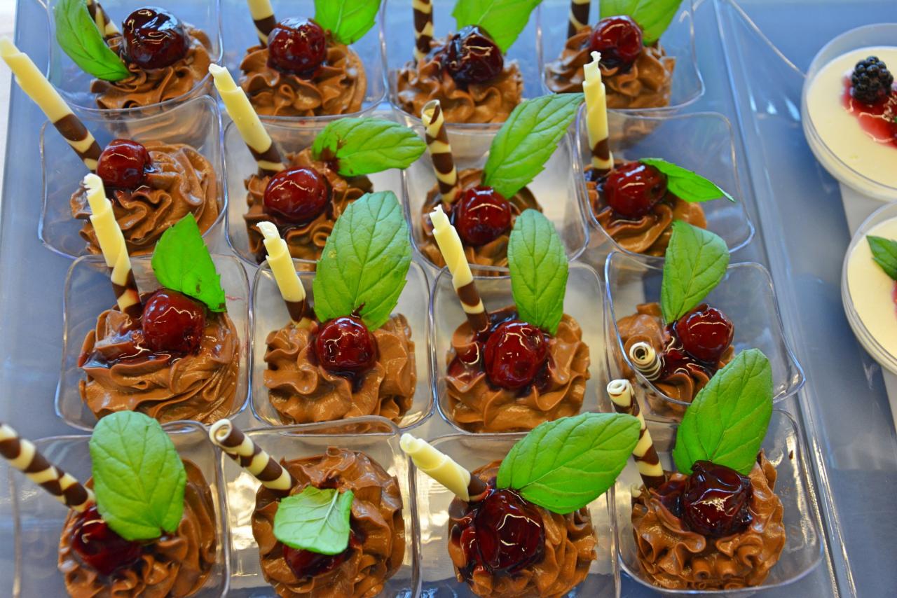 Mini deserki czekoladowe w przezroczystych pojemniczkach, udekorowane wiśnią, listkiem mięty i waflową rurką, ułożone na tacy.