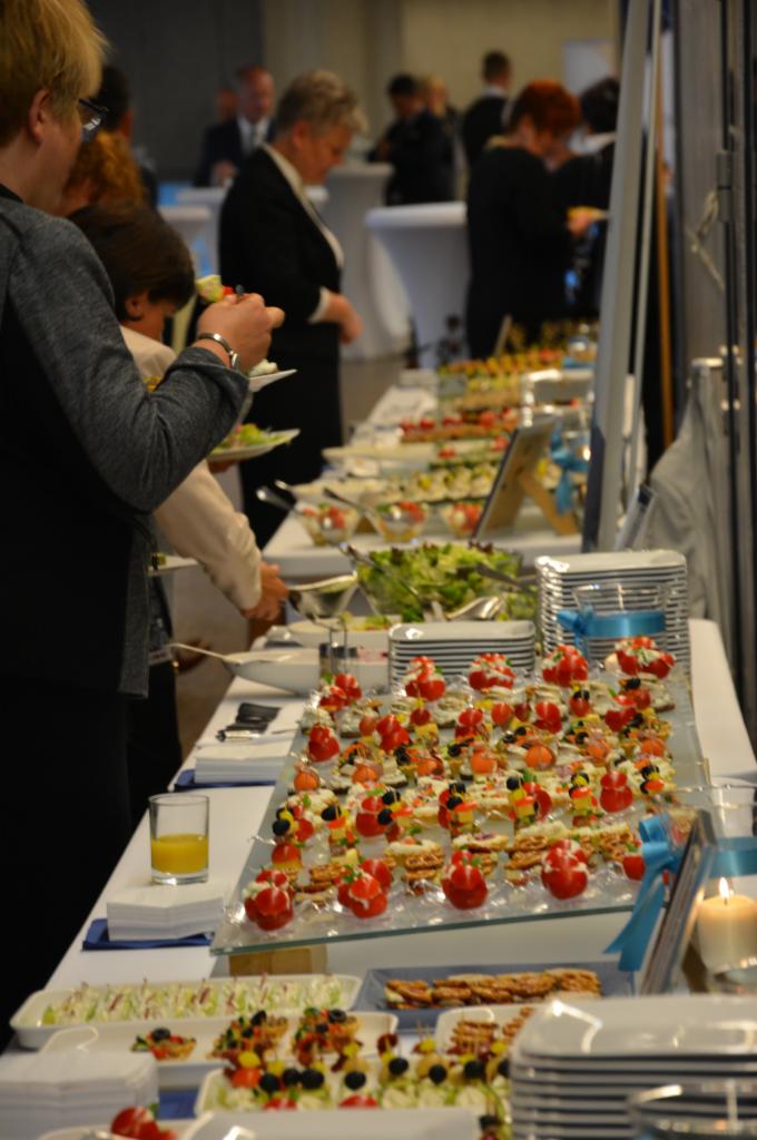 Bogato zastawiony stół z różnorodnymi przekąskami, sałatkami i deserami podczas przyjęcia, goście w tle wybierają jedzenie, świeca i szklanka soku na pierwszym planie.