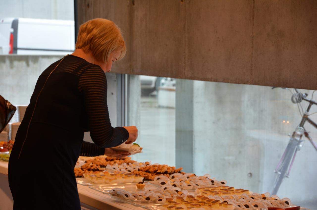 Kobieta w czarnej sukience nakłada przekąski na talerzyk z bufetu podczas wydarzenia, w tle widoczne okno i rower.