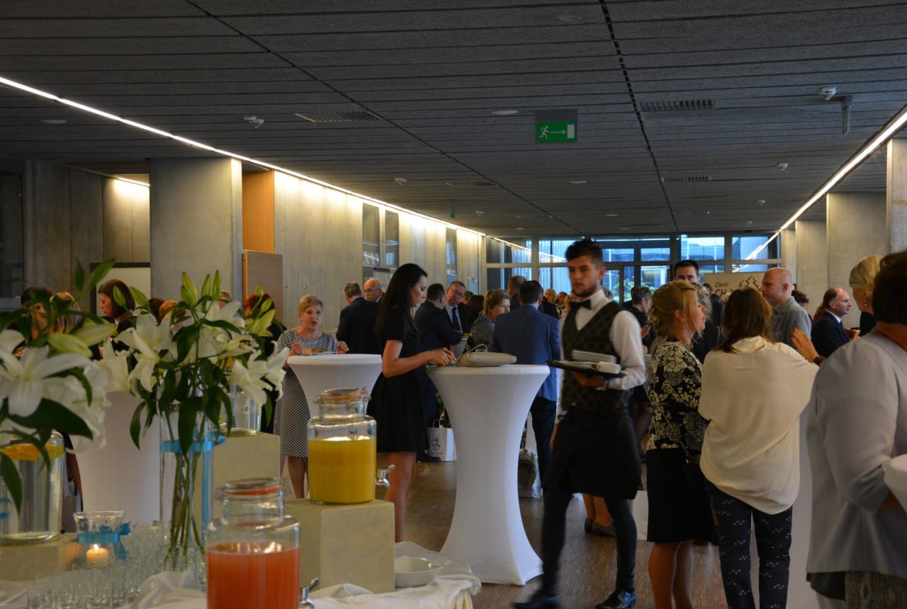 Eleganckie przyjęcie: kelner w muszce z tacą przechodzi obok gości stojących przy białych stolikach koktajlowych, na pierwszym planie szklane pojemniki z sokami i wazony z białymi liliami.