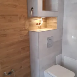 Nowoczesna łazienka w trakcie wykańczania: wisząca toaleta, drewnopodobne płytki na ścianie, podświetlana wnęka z wystającymi przewodami elektrycznymi, szara zabudowa.