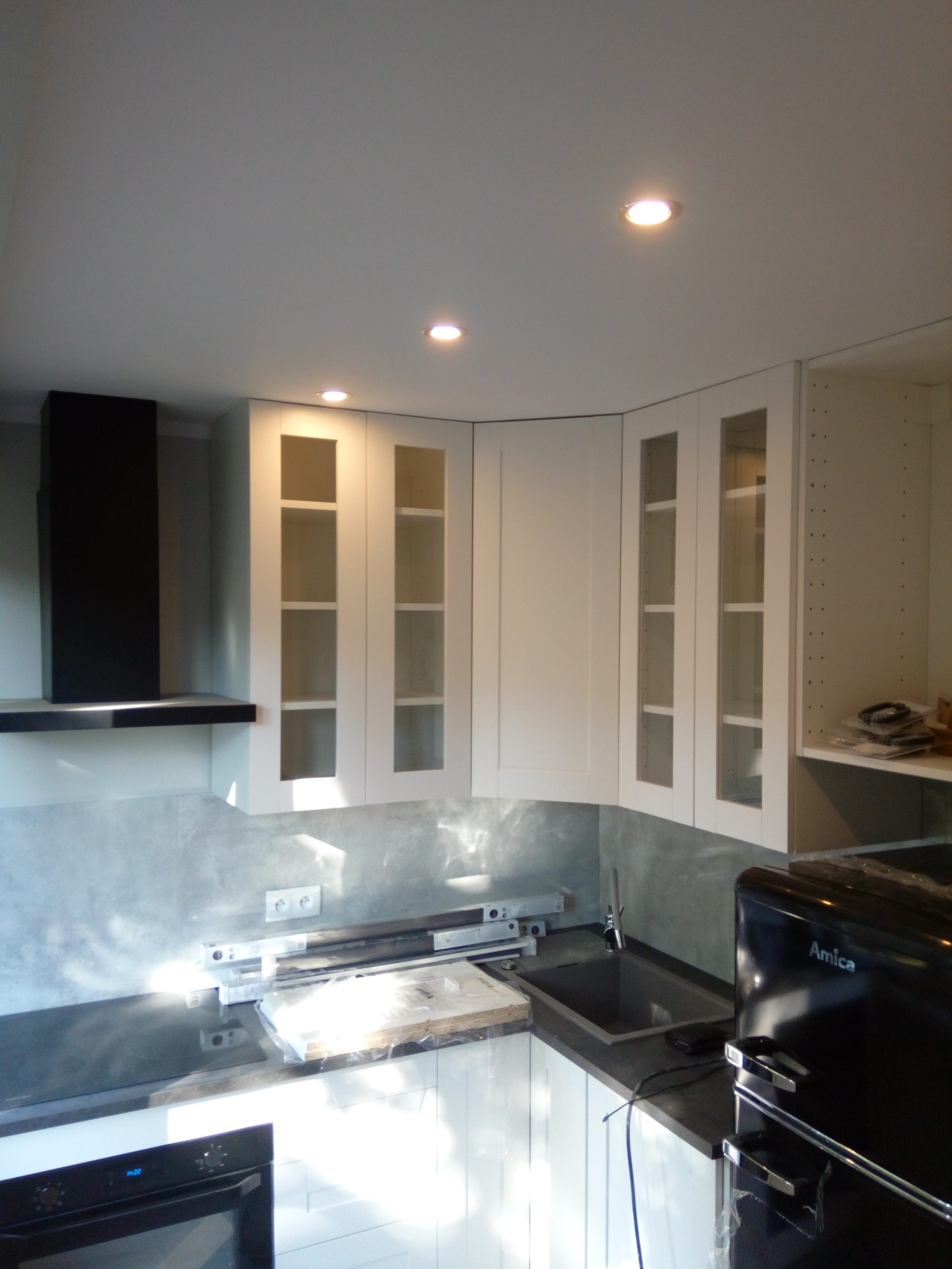 Wnętrze kuchni w trakcie montażu: białe szafki z przeszklonymi frontami, czarny okap, ciemny blat, czarna lodówka Amica, poziomice na blacie, surowe ściany między szafkami.