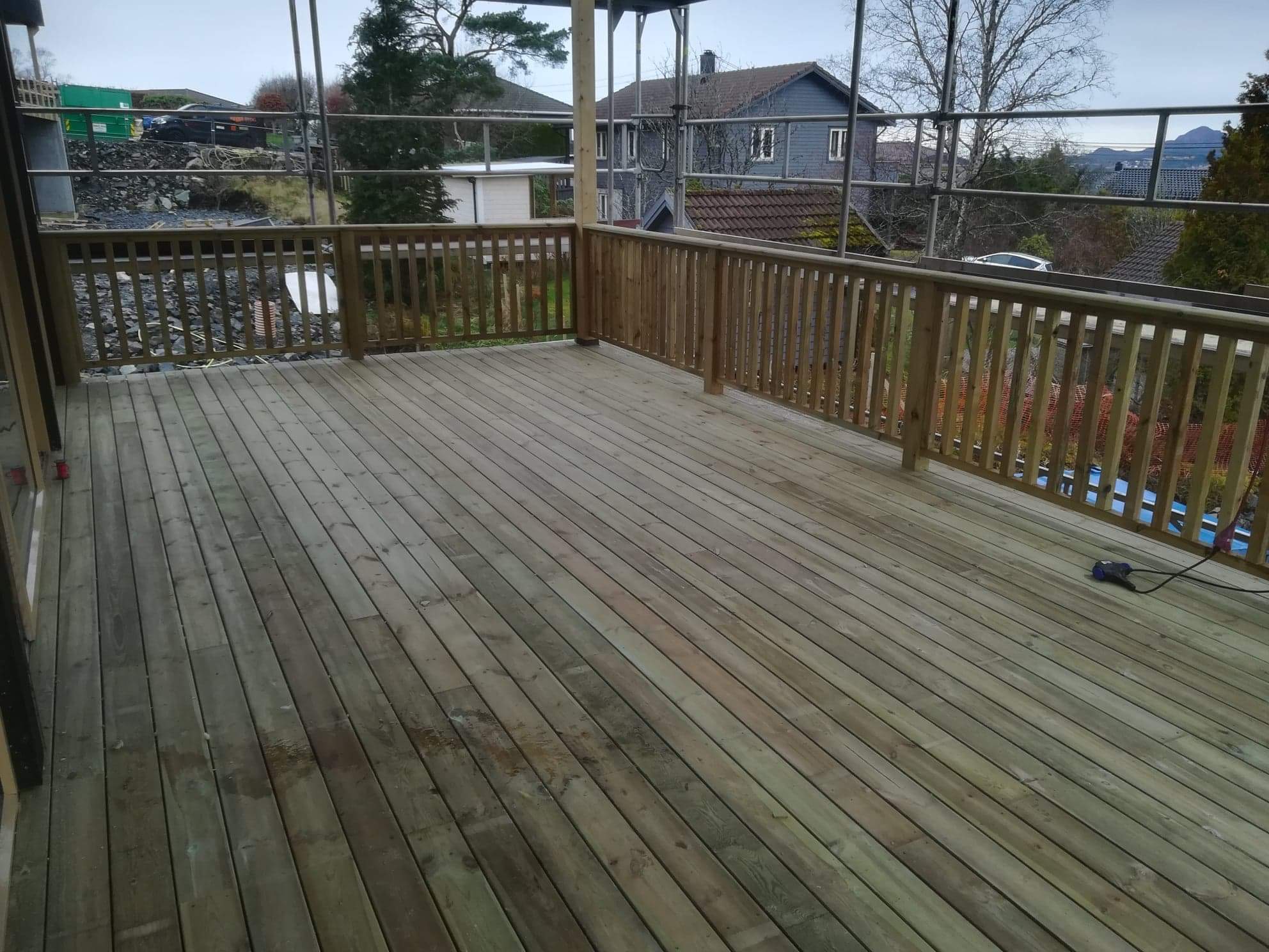 Drewniany taras z balustradą w trakcie budowy, widoczne rusztowania i narzędzie elektryczne na podłodze, domy w tle.