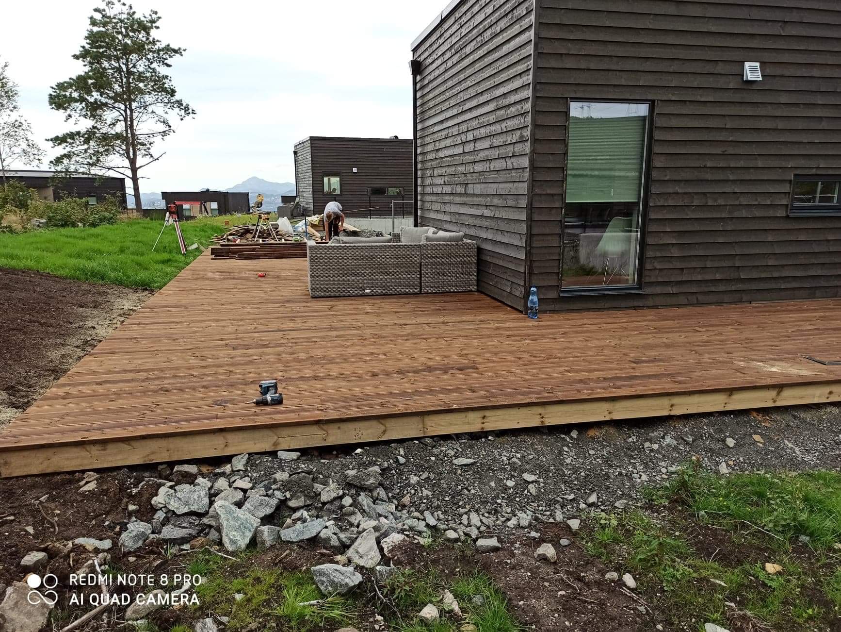 Brązowy drewniany taras przy ciemnym domu z desek, widoczne narzędzia i meble ogrodowe, w tle krajobraz z drzewami i wzgórzami.