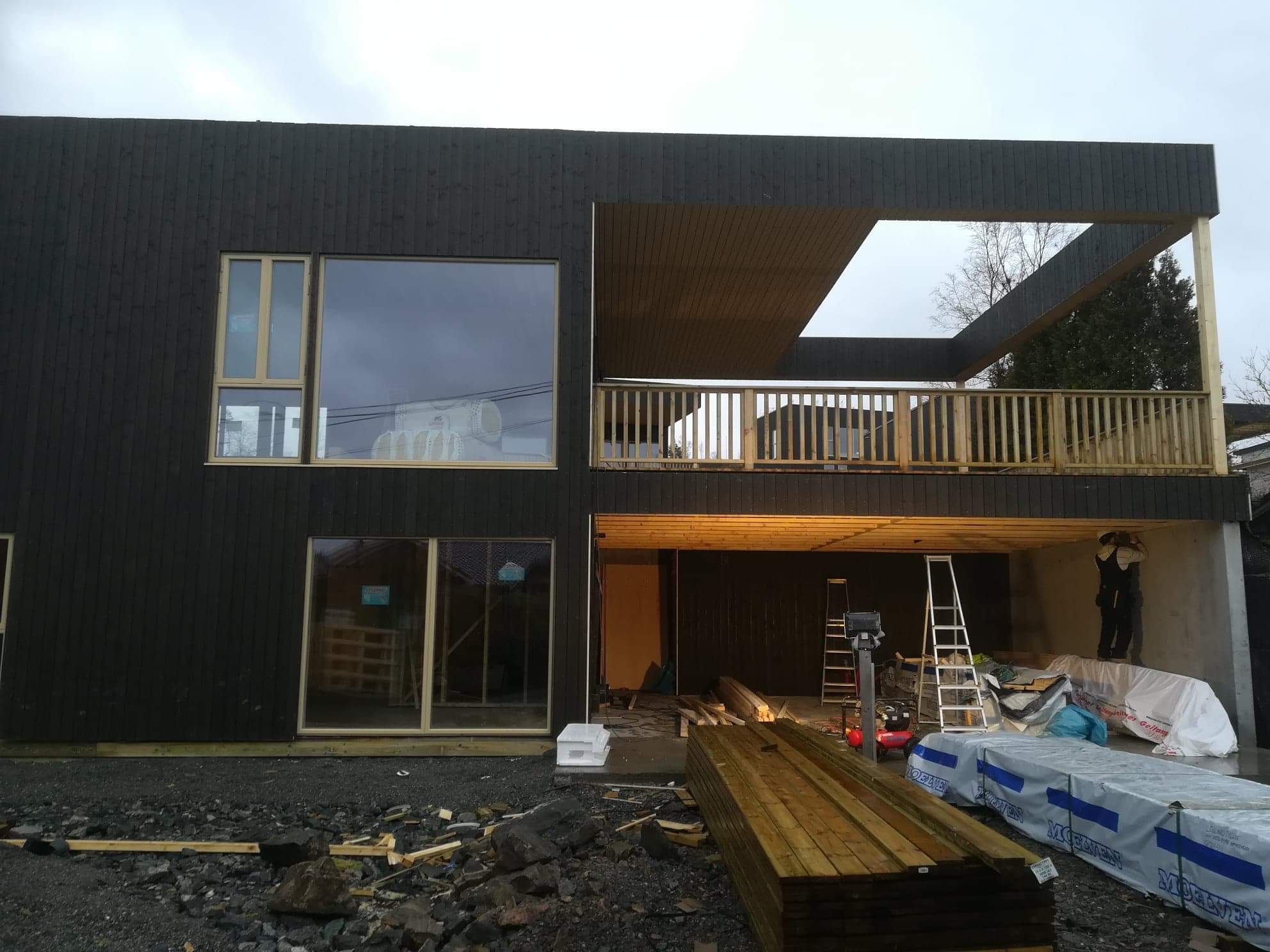 Nowoczesny, dwukondygnacyjny budynek w trakcie budowy, z czarną, drewnianą elewacją, dużymi oknami z jasnymi ramami i otwartym balkonem z drewnianą balustradą. Na placu budowy widoczne materiały...