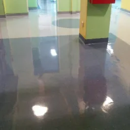 Błyszcząca, wielokolorowa podłoga w korytarzu biurowym, z widocznymi refleksami światła i przejściami kolorystycznymi między szarym, niebieskim i białym, otoczona ścianami w odcieniach zieleni...