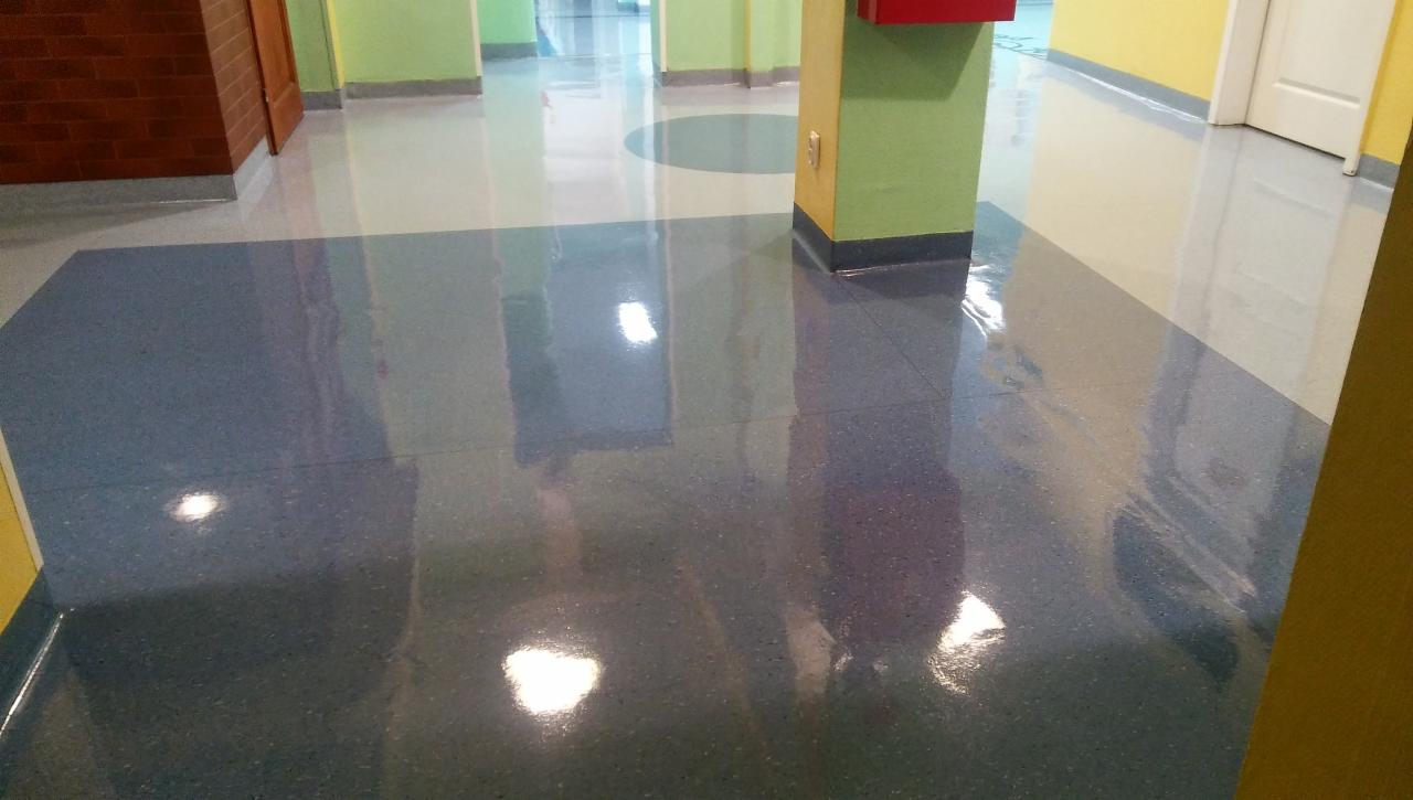 Błyszcząca, wielokolorowa podłoga w korytarzu biurowym, z widocznymi refleksami światła i przejściami kolorystycznymi między szarym, niebieskim i białym, otoczona ścianami w odcieniach zieleni...