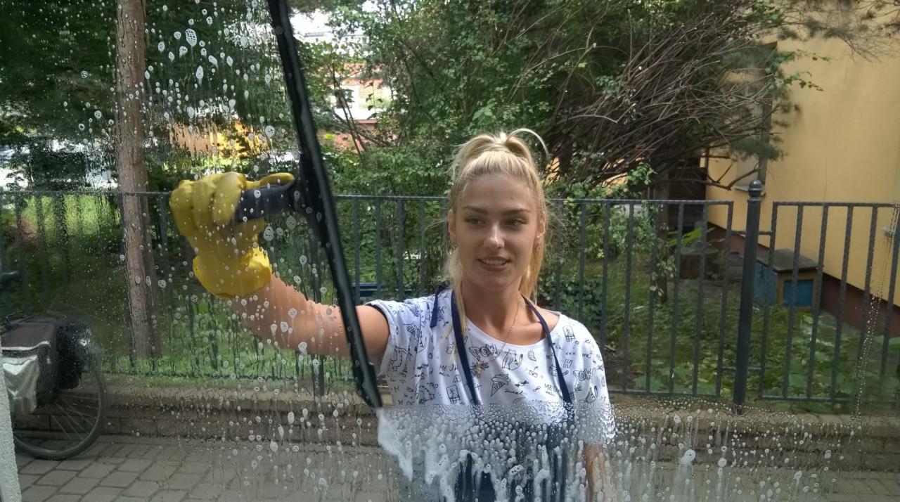 Uśmiechnięta kobieta w żółtych rękawicach myje okno za pomocą ściągaczki, widoczne krople wody i piana, w tle zieleń i budynek.