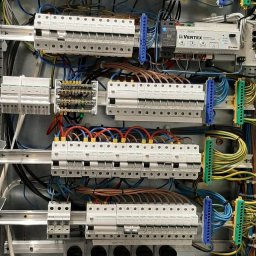 Eko-Instalacje - Rozdzielnica elektryczna z wieloma bezpiecznikami, przewodami o różnych kolorach, listwami zaciskowymi i modułem Vertex, w metalowej obudowie, widok z bliska.