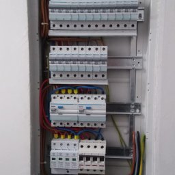Eko-Instalacje - Otwarta skrzynka elektryczna wnękowa z widocznymi bezpiecznikami, przewodami o różnych kolorach i listwami zaciskowymi, zamontowana w białej ścianie.