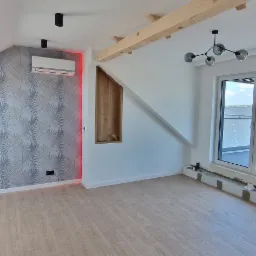 Wnętrze poddasza z drewnianą podłogą, widocznym klimatyzatorem z czerwonym podświetleniem, drewnianą wnęką w ścianie i surowymi belkami stropowymi.