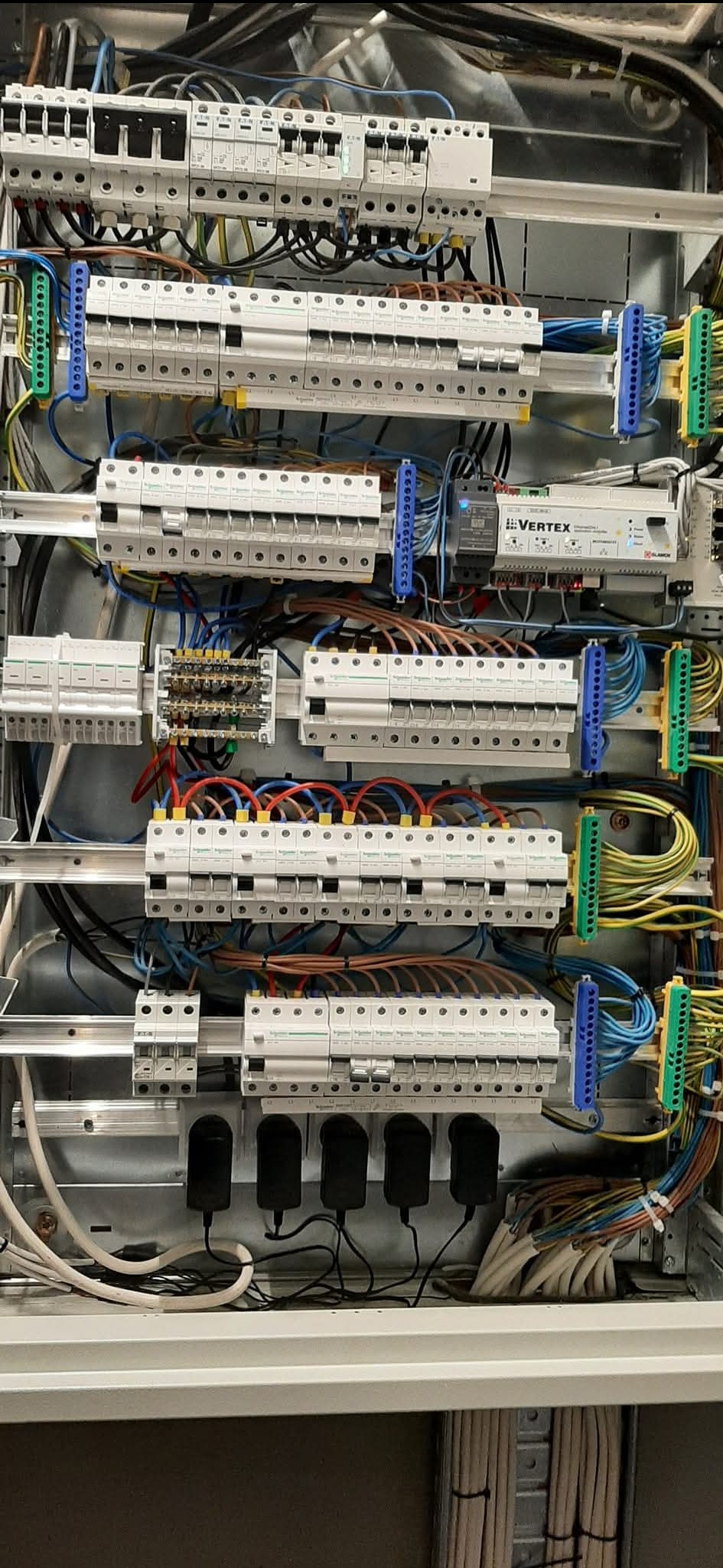 Rozdzielnica elektryczna z wieloma bezpiecznikami, przewodami o różnych kolorach, listwami zaciskowymi i modułem Vertex, w metalowej obudowie, widok z bliska.