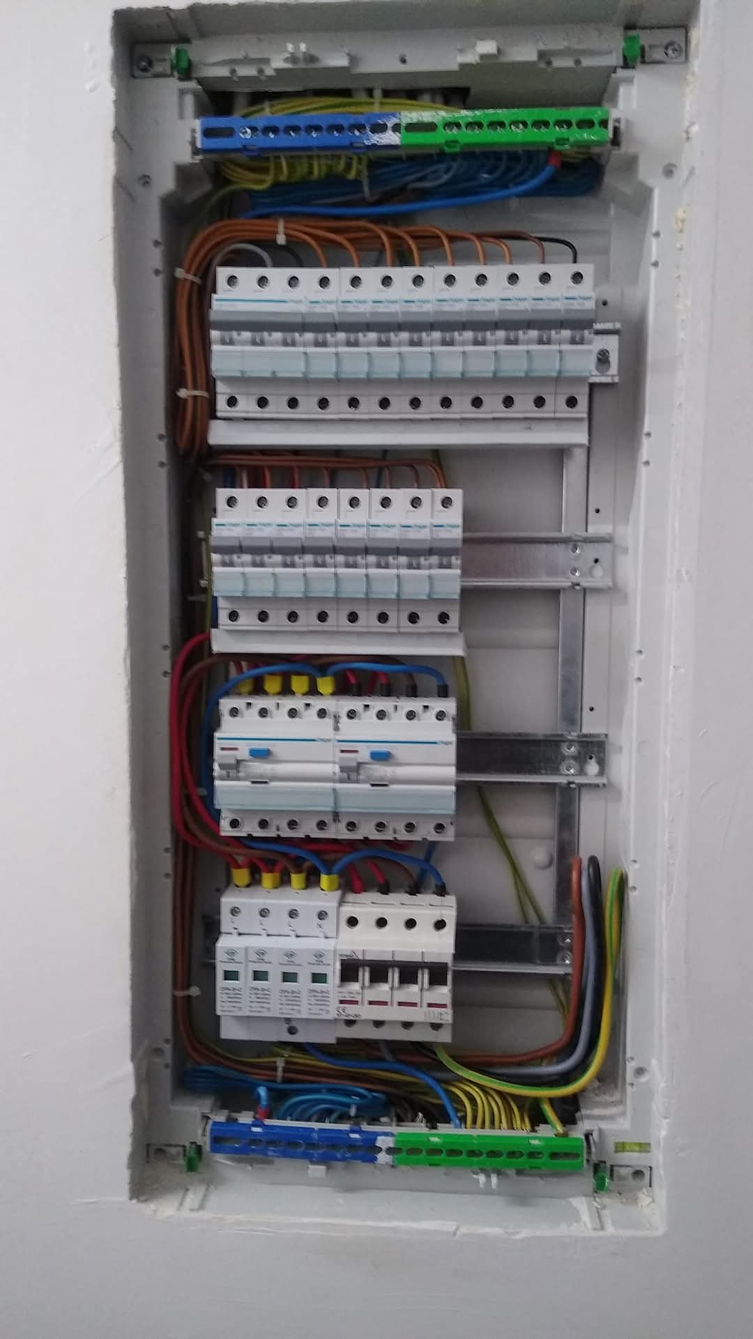 Otwarta skrzynka elektryczna wnękowa z widocznymi bezpiecznikami, przewodami o różnych kolorach i listwami zaciskowymi, zamontowana w białej ścianie.