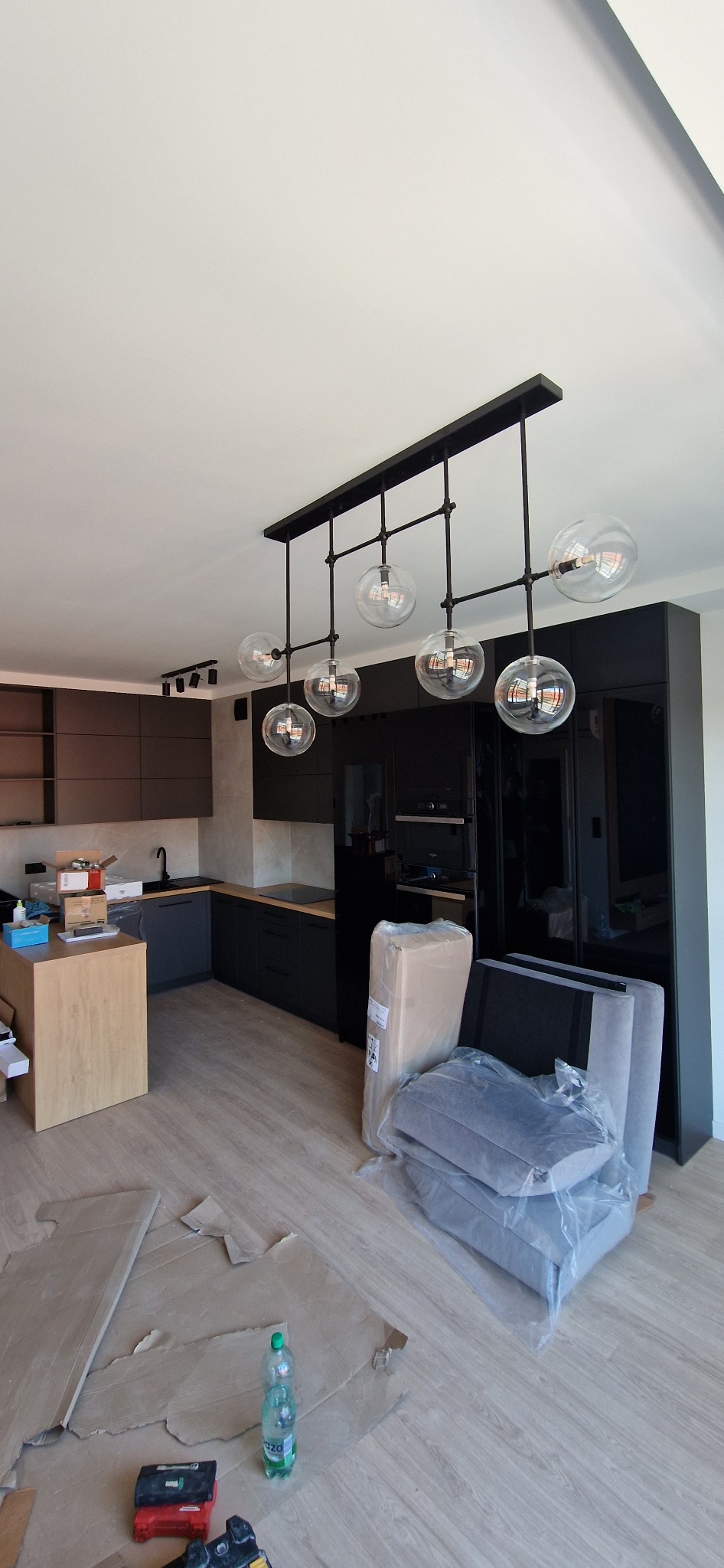 Nowoczesna kuchnia w trakcie wykańczania z zawieszonym czarnym żyrandolem z sześcioma szklanymi kulami, widoczne kartony po meblach i narzędzia na podłodze.