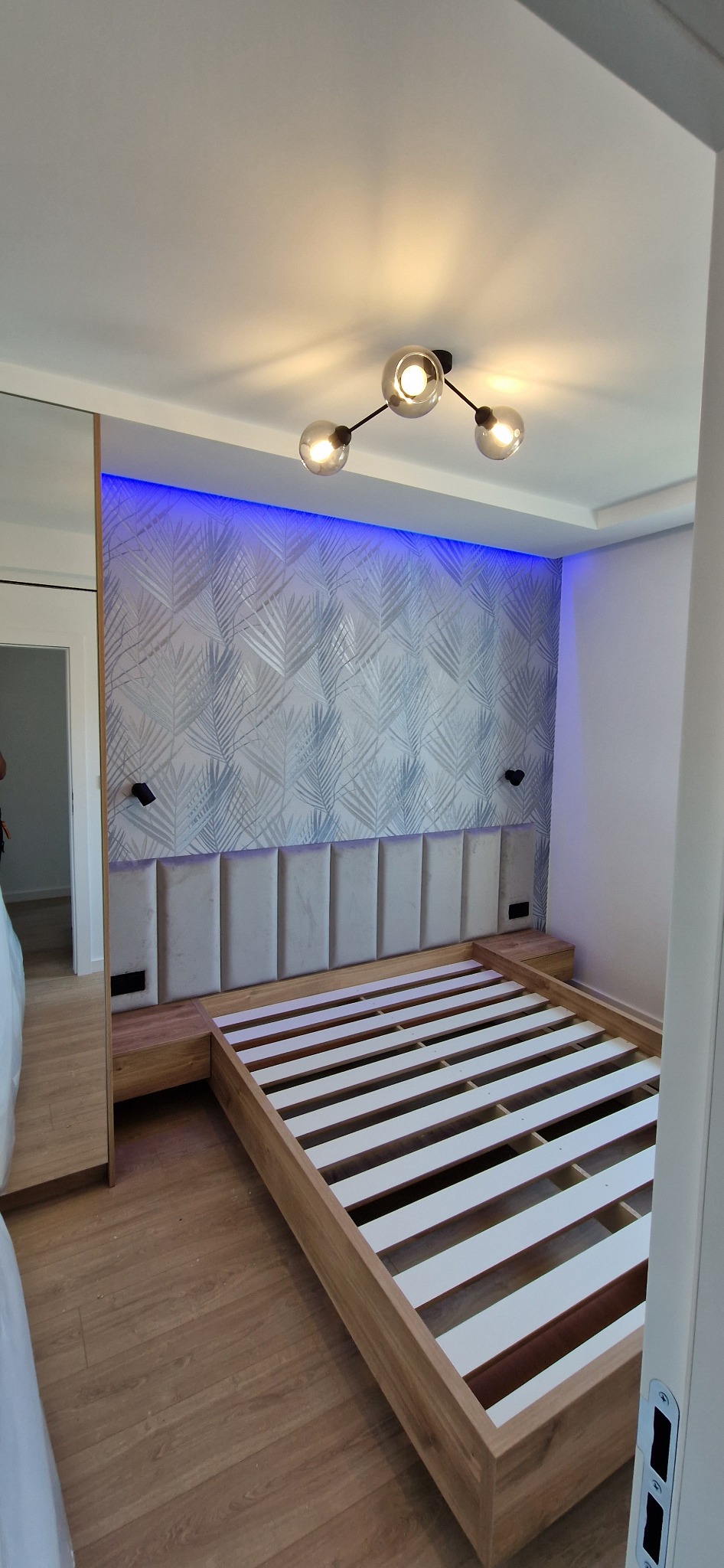 Nowoczesna sypialnia z drewnianą ramą łóżka, tapicerowanym wezgłowiem w kolorze ecru, podświetleniem LED w kolorze niebieskim za wezgłowiem i lampą sufitową z trzema szklanymi kloszami.