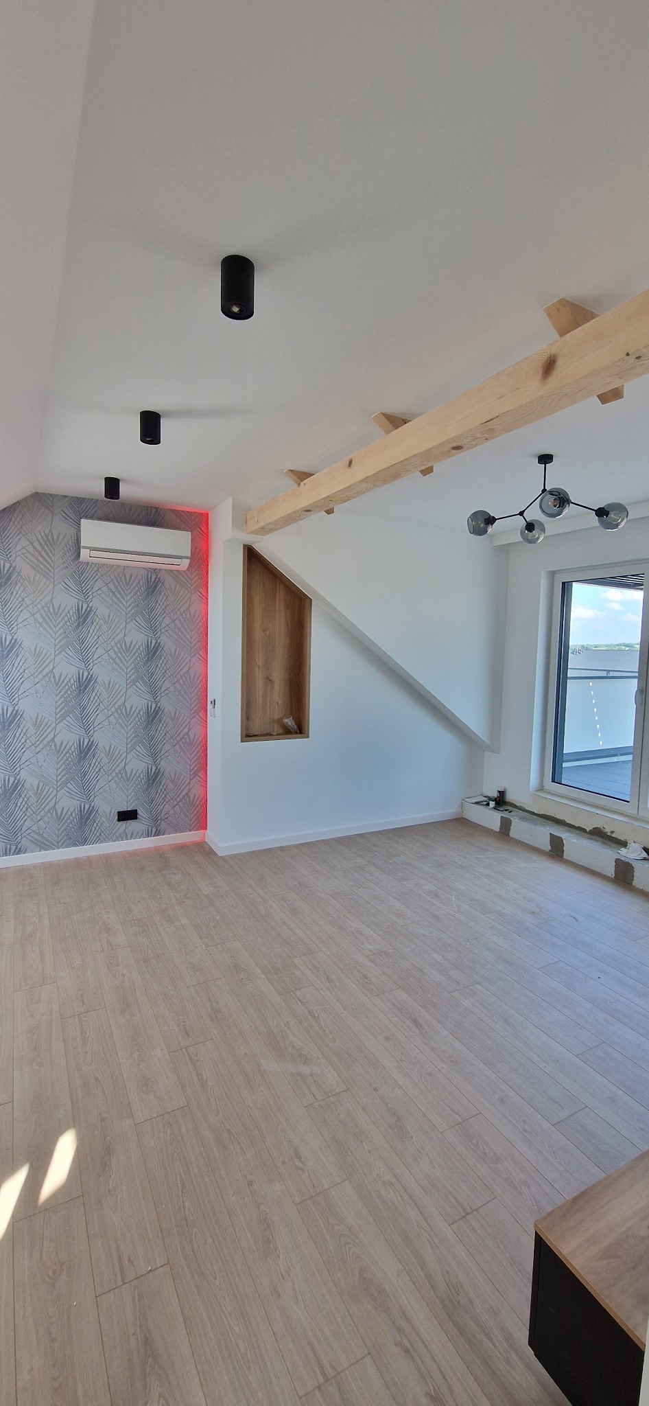 Wnętrze poddasza z drewnianą podłogą, widocznym klimatyzatorem z czerwonym podświetleniem, drewnianą wnęką w ścianie i surowymi belkami stropowymi.