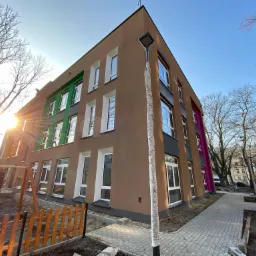 Nowo wybudowany, trzypiętrowy budynek z brązową elewacją, fragment placu zabaw z huśtawką i drewnianym ogrodzeniem, chodnik z kostki brukowej, lampa owinięta folią ochronną, w tle drzewa bez liści...