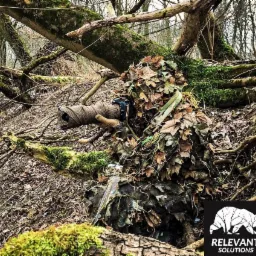 Osoba w kamuflażu leśnym typu ghillie suit, trzymająca obiektyw aparatu owinięty materiałem maskującym, ukryta w zaroślach na tle leśnym.