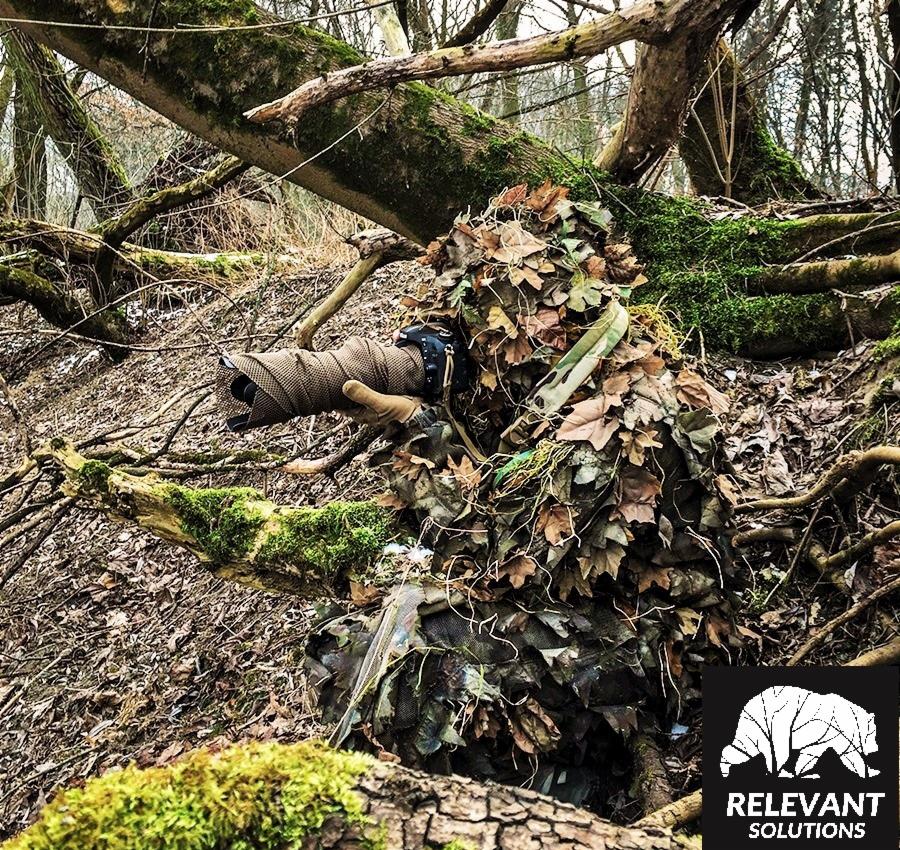 Osoba w kamuflażu leśnym typu ghillie suit, trzymająca obiektyw aparatu owinięty materiałem maskującym, ukryta w zaroślach na tle leśnym.
