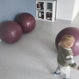 Jasne pomieszczenie z trzema dużymi, bordowymi piłkami do ćwiczeń, białym regałem z książkami i rozmazanym małym dzieckiem w ruchu.