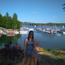 Uśmiechnięta kobieta w okularach przeciwsłonecznych siedzi na ławce na tle przystani z jachtami i zielenią.