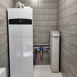 Biała, pionowa jednostka grzewcza Ariston z wyświetlaczem, rurami i filtrem wody w pomieszczeniu wyłożonym szarymi płytkami.