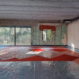 Czerwone rury ogrzewania podłogowego rozłożone na folii z nadrukiem siatki w surowym wnętrzu z dużymi oknami i betonowym sufitem.