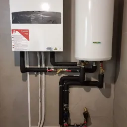 Biały piec gazowy Ariston zawieszony obok białego zasobnika ciepłej wody, połączone czarnymi rurami z zaworami na szarych płytkach ściennych.