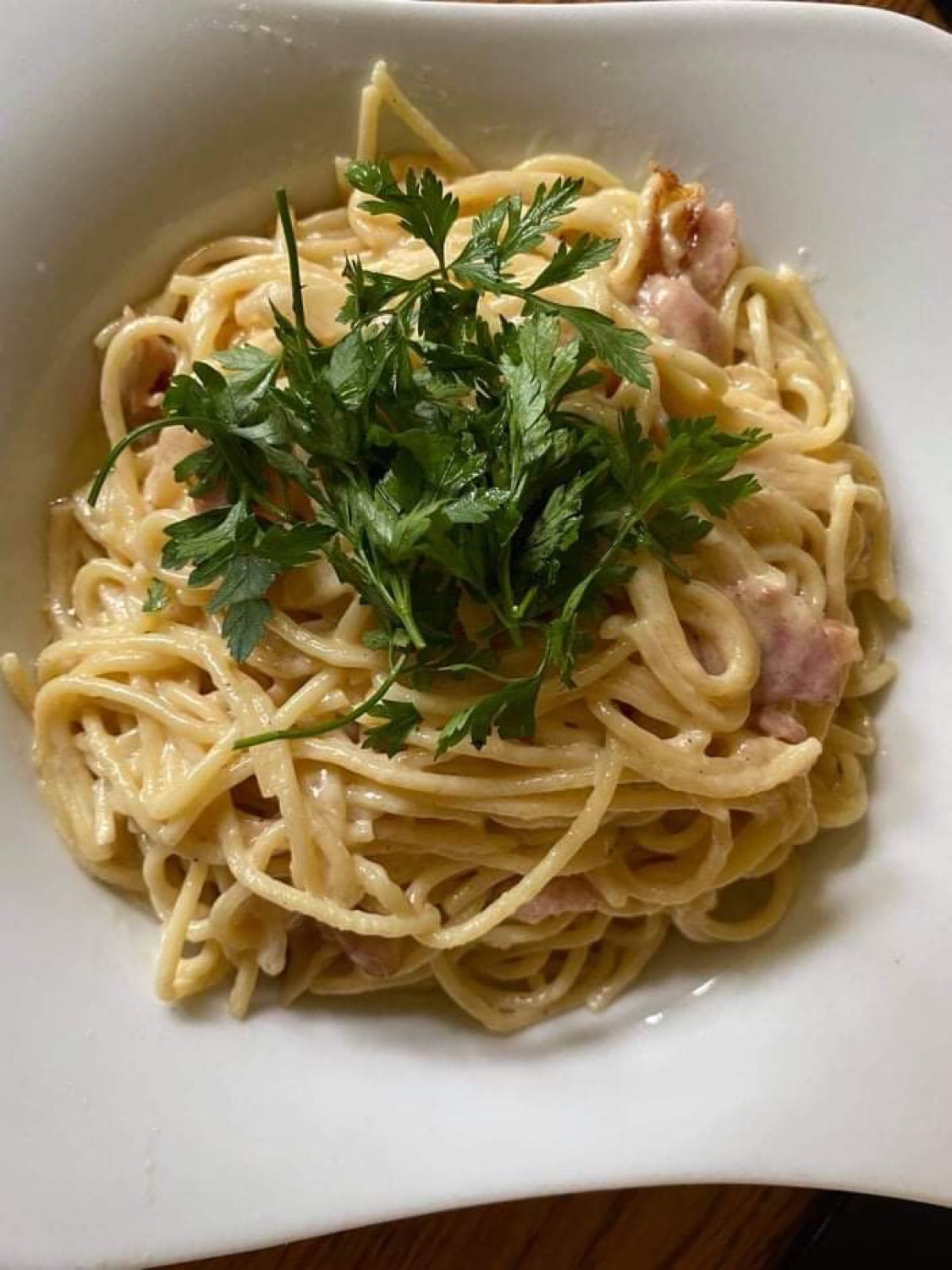 Kremowe spaghetti carbonara z boczkiem i świeżą natką pietruszki w białej, kwadratowej misce, widok z góry.