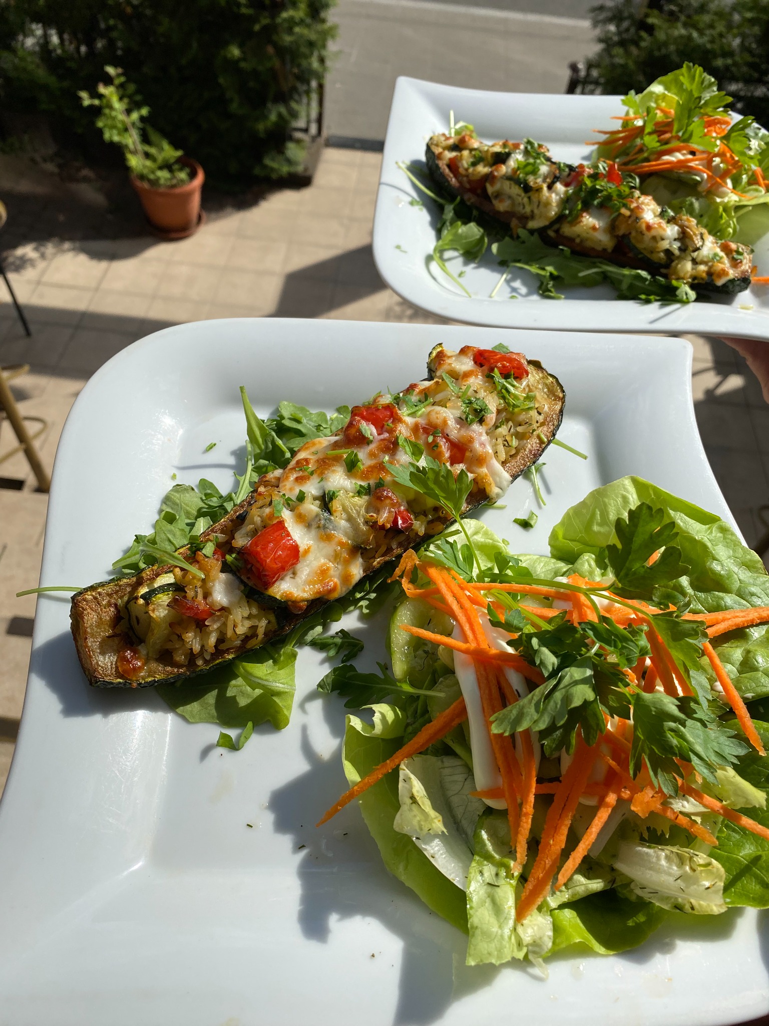 Dwa talerze z pieczoną cukinią faszerowaną ryżem i warzywami, zapieczoną serem, podane z sałatką z marchewki i zielonych liści, ustawione na zewnątrz w słoneczny dzień.