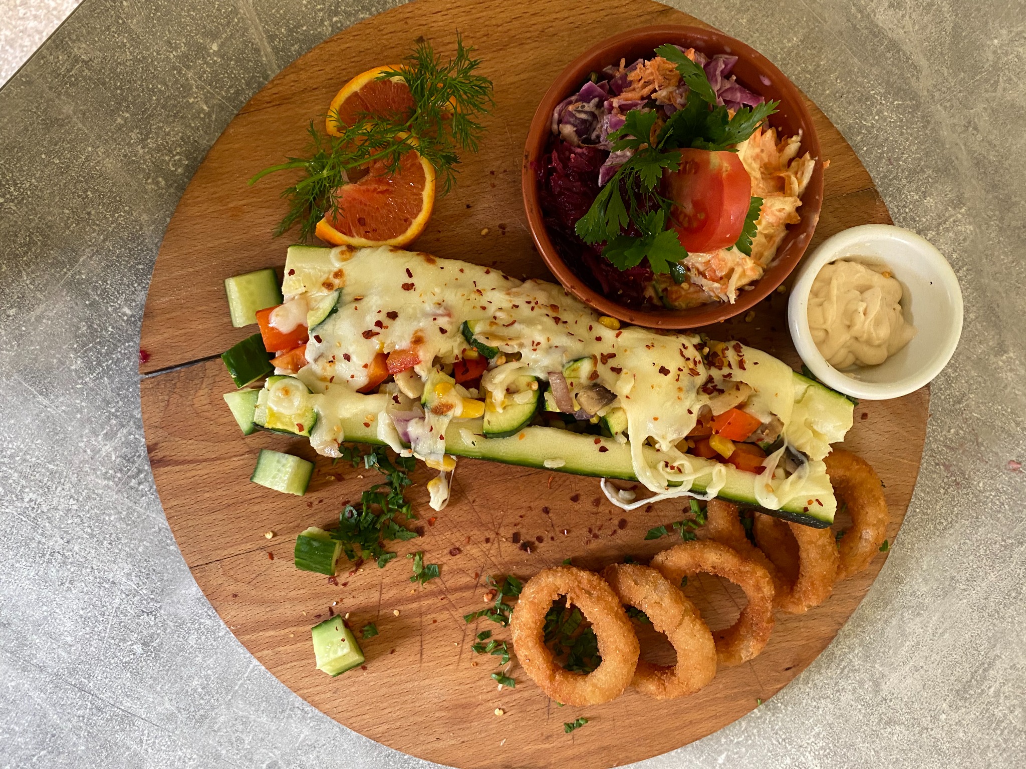 Pieczona cukinia faszerowana warzywami pod serową pierzynką, podana z sałatką warzywną, sosem i krążkami cebulowymi na drewnianej desce.