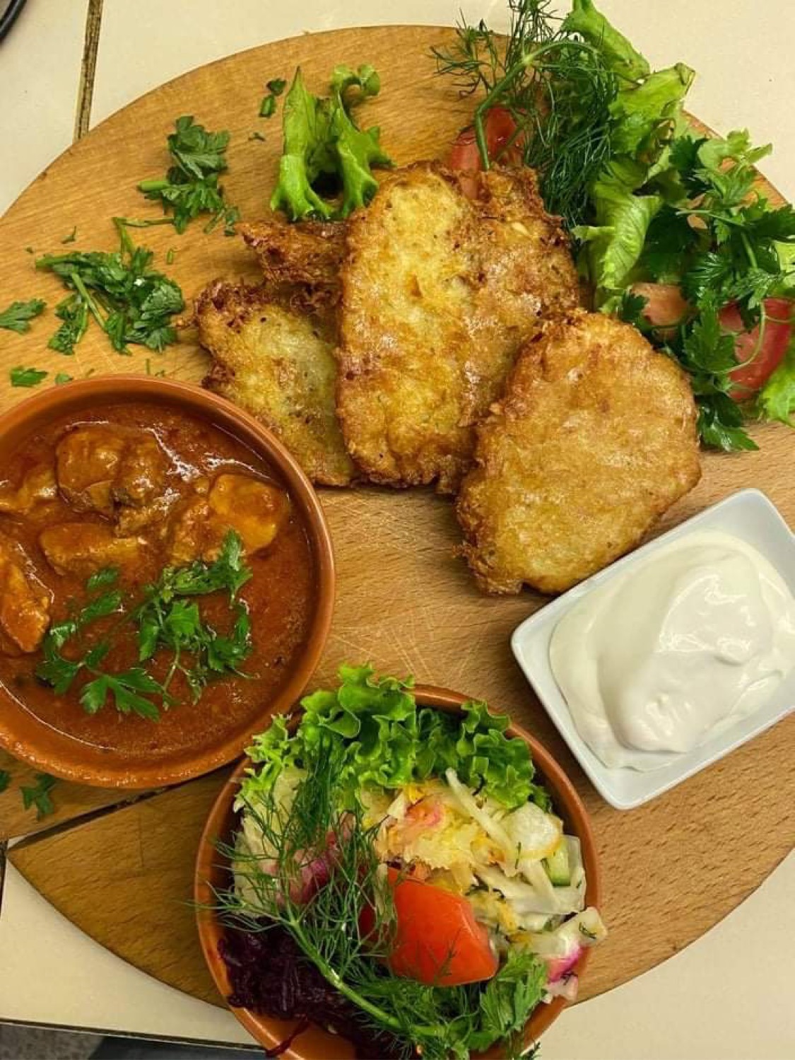 Drewniana deska z plackami ziemniaczanymi, gulaszem, surówką i sosem śmietanowym, udekorowana zieleniną.