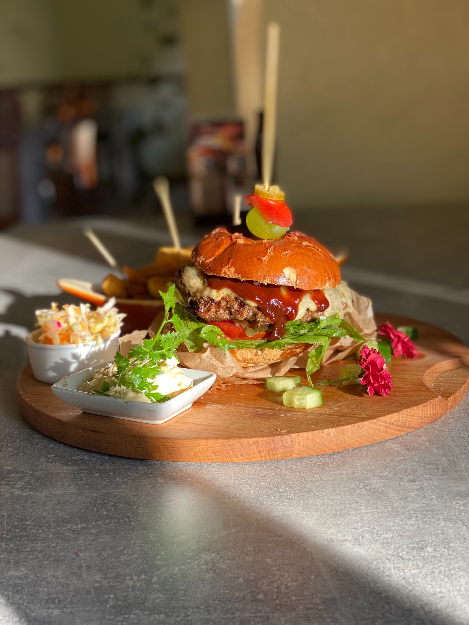 Apetyczny burger z sosem, warzywami i serem, podany z frytkami i sałatkami na drewnianej desce, udekorowany kwiatami i szaszłykiem z winogrona i papryki, oświetlony ciepłym światłem.