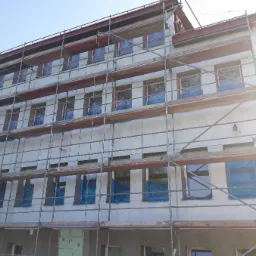 Budynek wielorodzinny w trakcie ocieplania, z oknami zabezpieczonymi folią i rusztowaniem, robotnicy pracują na rusztowaniu.