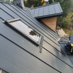 Montaż pokrycia z blachy na dachu z oknem dachowym i kominem, widoczne drewniane elementy konstrukcji dachu i fragment rusztowania.