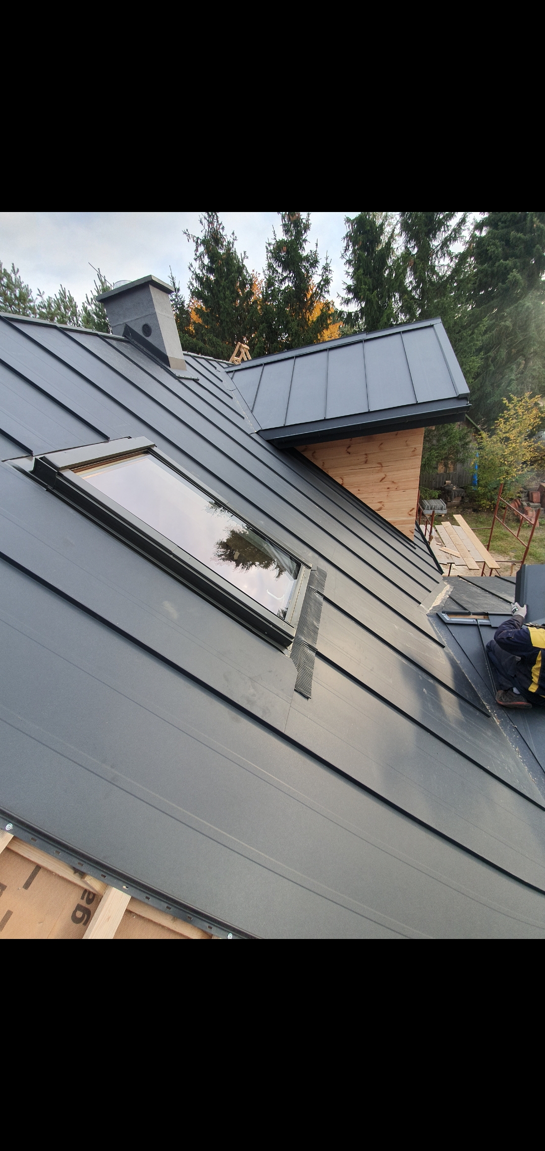 Montaż pokrycia z blachy na dachu z oknem dachowym i kominem, widoczne drewniane elementy konstrukcji dachu i fragment rusztowania.