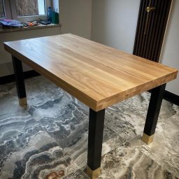 P.H.U. Ja-Ry Jacek Rycharski - Drewniany stół z blatem z jasnego drewna i czarnymi nogami z mosiężnymi detalami, ustawiony na marmurowej podłodze. Widoczne okno i dekoracyjny panel ścienny.