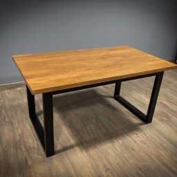 P.H.U. Ja-Ry Jacek Rycharski - Stół z blatem z jasnego drewna i czarnymi, metalowymi nogami w kształcie trapezu. Minimalistyczny design, widoczne usłojenie drewna, szare tło i drewniana podłoga.