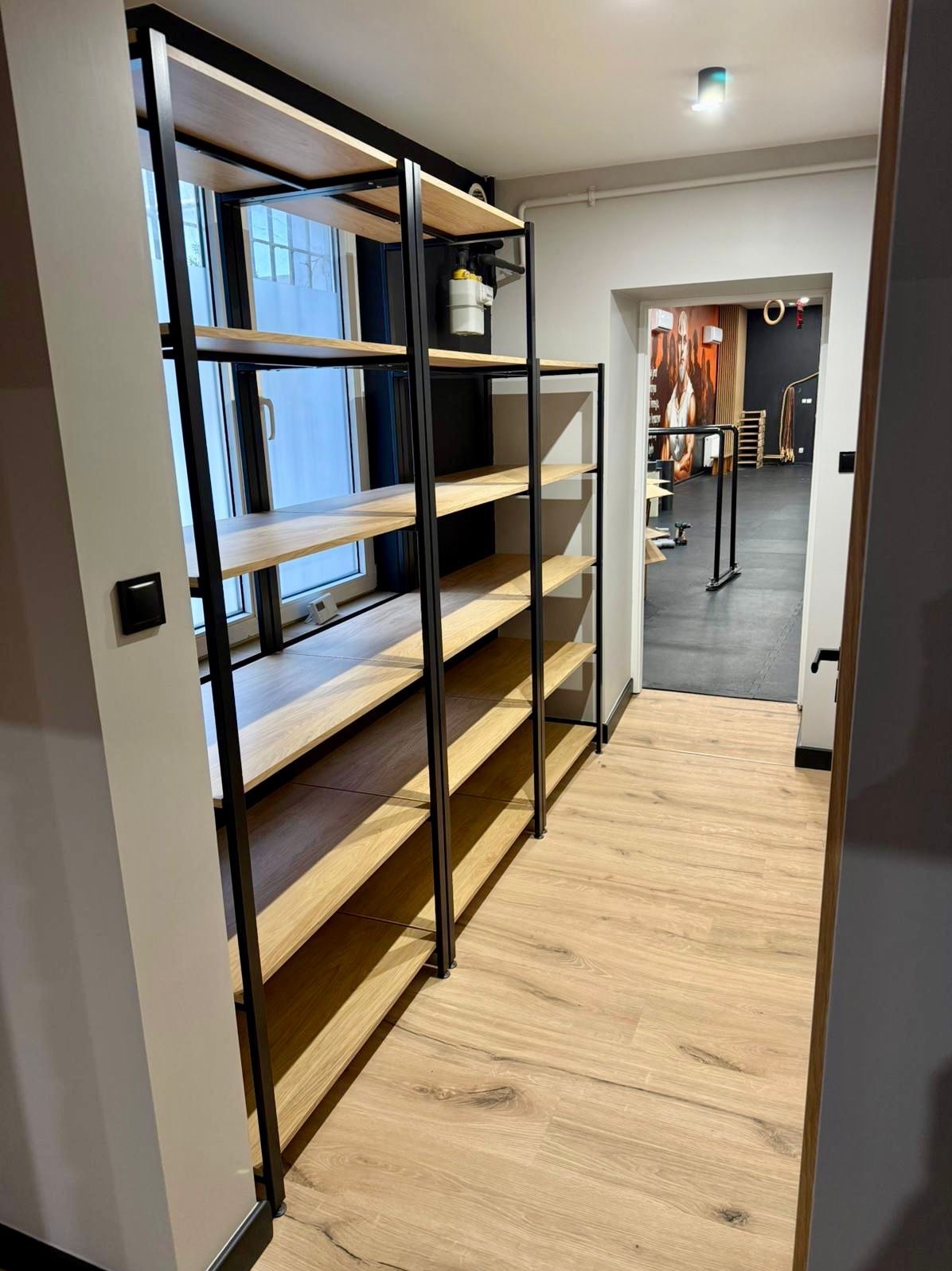 Nowoczesny regał z drewnianymi półkami i czarną metalową konstrukcją, umieszczony w pomieszczeniu z widokiem na okno i salę ćwiczeń w tle.