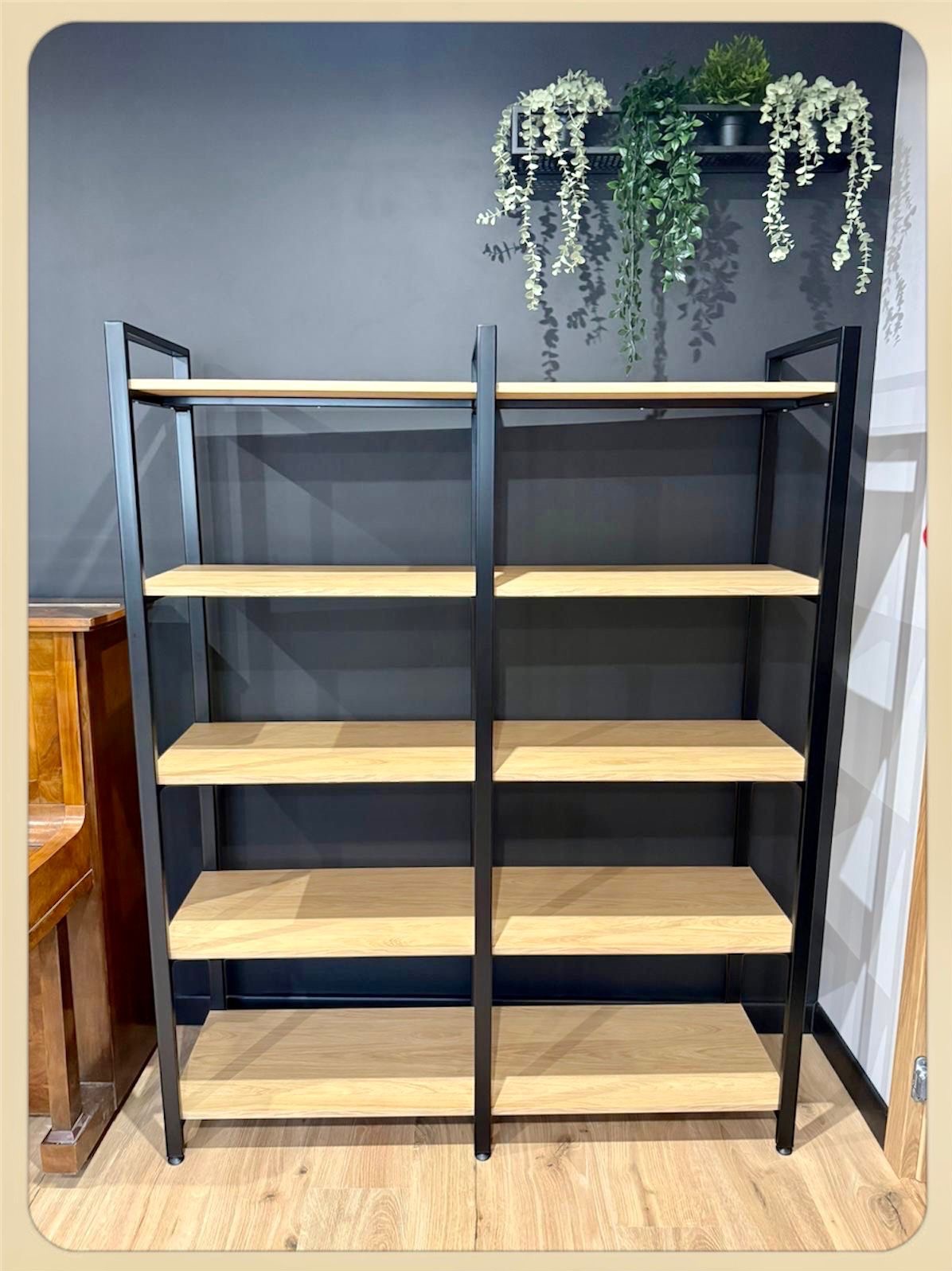 Regał z drewnianymi półkami i czarną metalową ramą, stojący na drewnianej podłodze, z roślinami wiszącymi na ścianie w tle. Minimalistyczny design.