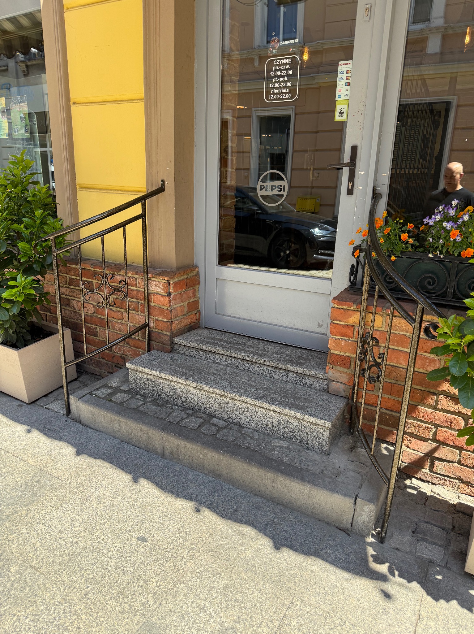 Metalowa balustrada schodowa z ozdobnymi elementami, zamontowana przy wejściu do lokalu z ceglanym wykończeniem. Widoczne schody z szarego kamienia i rośliny w donicach.