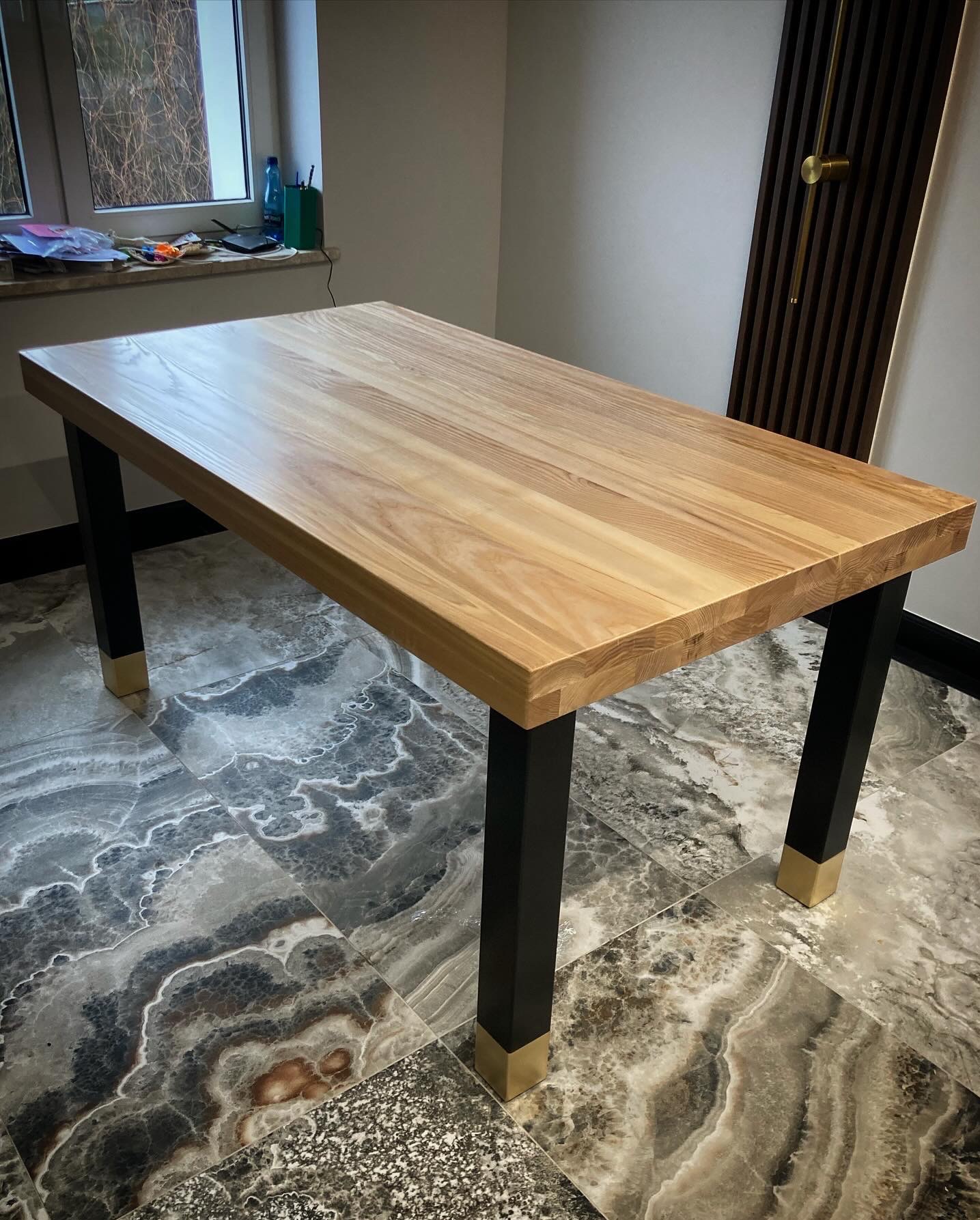 Drewniany stół z blatem z jasnego drewna i czarnymi nogami z mosiężnymi detalami, ustawiony na marmurowej podłodze. Widoczne okno i dekoracyjny panel ścienny.