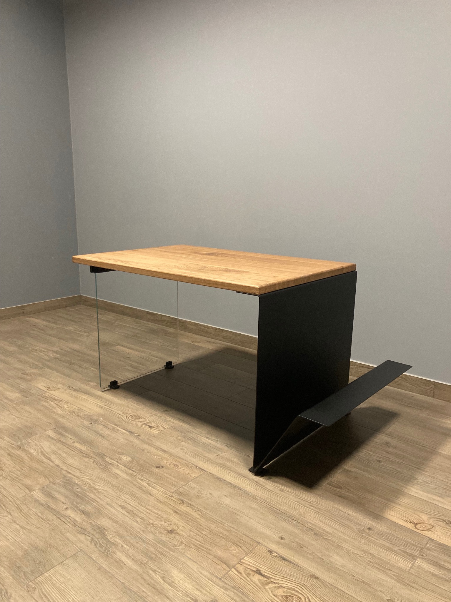 Nowoczesny stół z blatem z jasnego drewna, szklanymi nogami i czarną, metalową podstawą z elementem na gazety. Minimalistyczny design, neutralne tło.