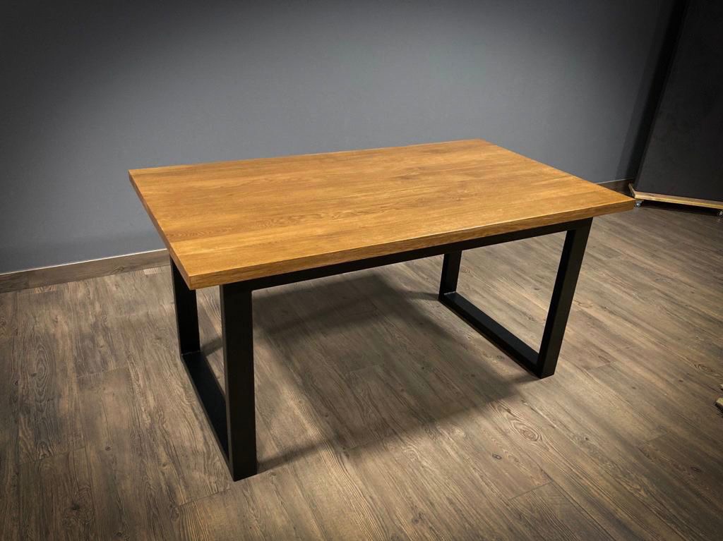 Stół z blatem z jasnego drewna i czarnymi, metalowymi nogami w kształcie trapezu. Minimalistyczny design, widoczne usłojenie drewna, szare tło i drewniana podłoga.