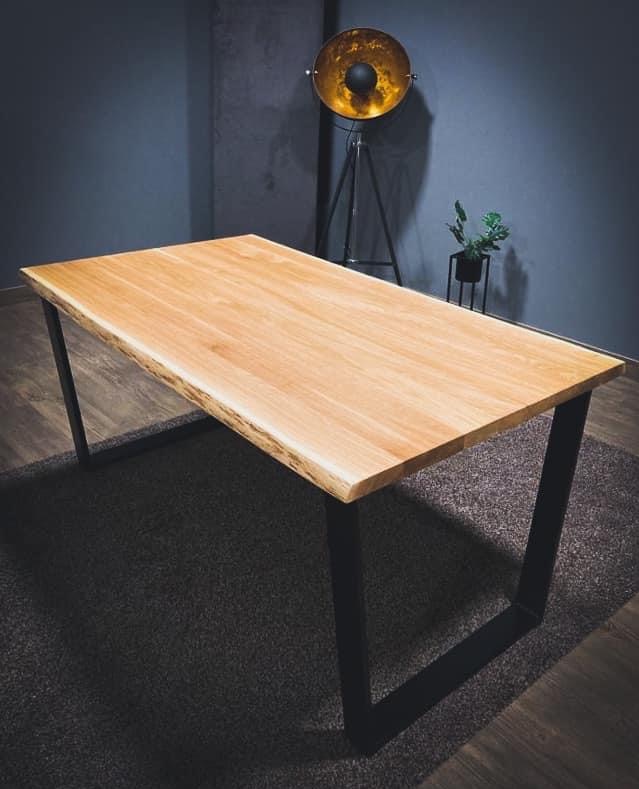 Stół z blatem z jasnego drewna o nieregularnych krawędziach, wsparty na czarnych, metalowych nogach w kształcie litery U. W tle lampa studyjna i roślina doniczkowa.