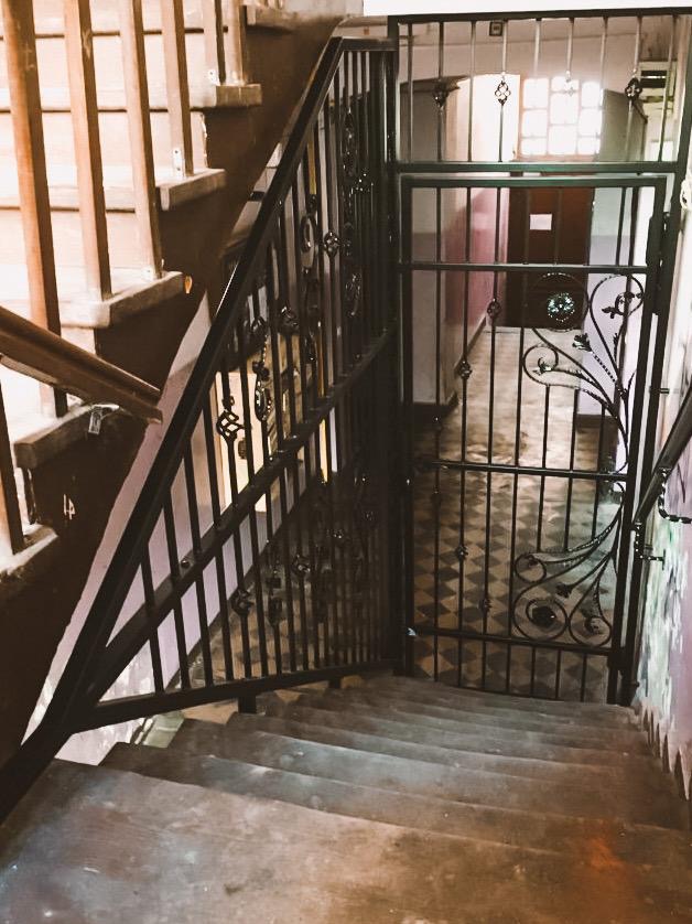 Kuta, czarna barierka schodowa z dekoracyjnymi elementami, zabezpieczająca wejście na klatkę schodową. Widok z góry na schody z szarego betonu.