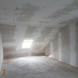 RiW Remonty i Wykończenia - Poddasze w trakcie remontu: ściany i sufit pokryte wstępną warstwą szpachli, widoczne kable elektryczne wystające ze ścian, okno dachowe w skosie, butelka z napojem na podłodze.